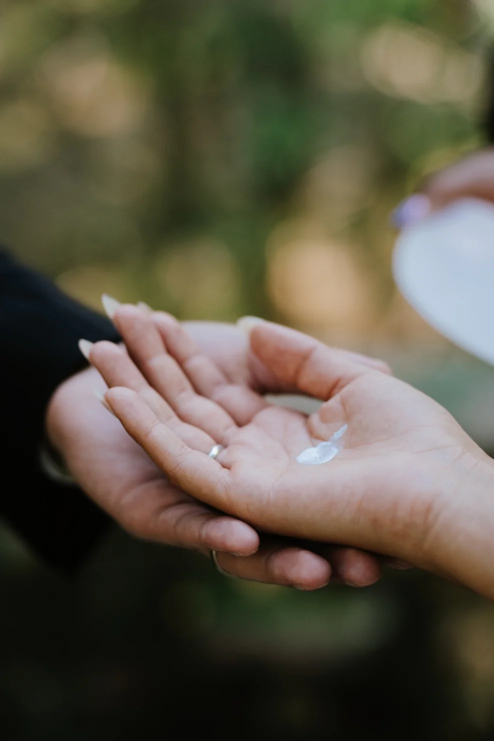 Gros plan sur deux mains entrelacées tenant délicatement une petite perle blanche sur un fond naturel flou.