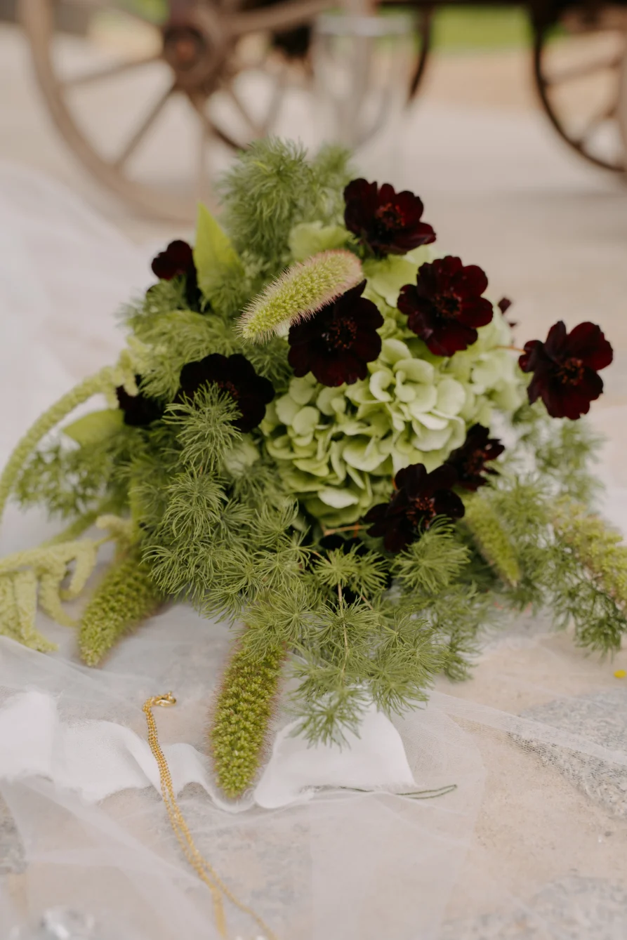 Bouquet champêtre aux fleurs bordeaux et vert tendre posé sur un voile blanc vaporeux.