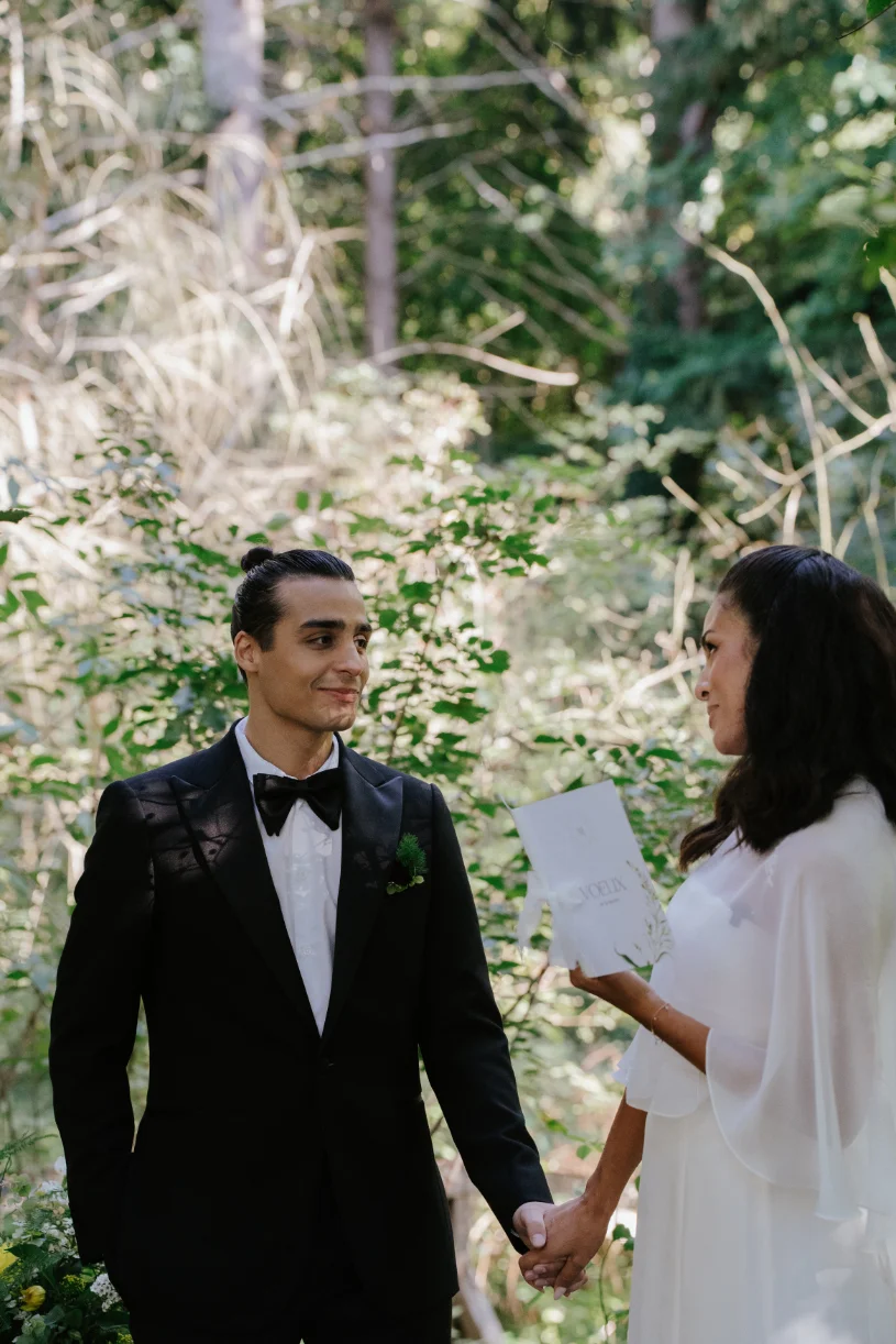 Un couple élégant se tient la main dans un sous-bois lumineux, le marié en smoking noir et la mariée en robe blanche.