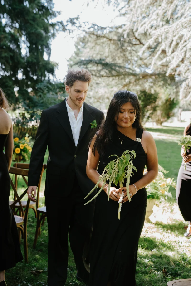 Un couple élégant en tenue noire pose dans un jardin ensoleillé, la femme tenant un bouquet de fleurs sauvages.