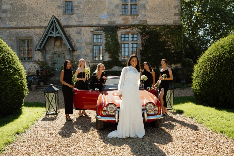 Une mariée en robe blanche et ses demoiselles d'honneur en noir posent devant une voiture rouge vintage et un manoir.