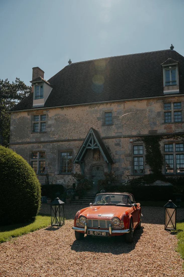 Voiture de collection rouge garée devant un manoir ancien sous un soleil éclatant, ambiance romantique et intemporelle.