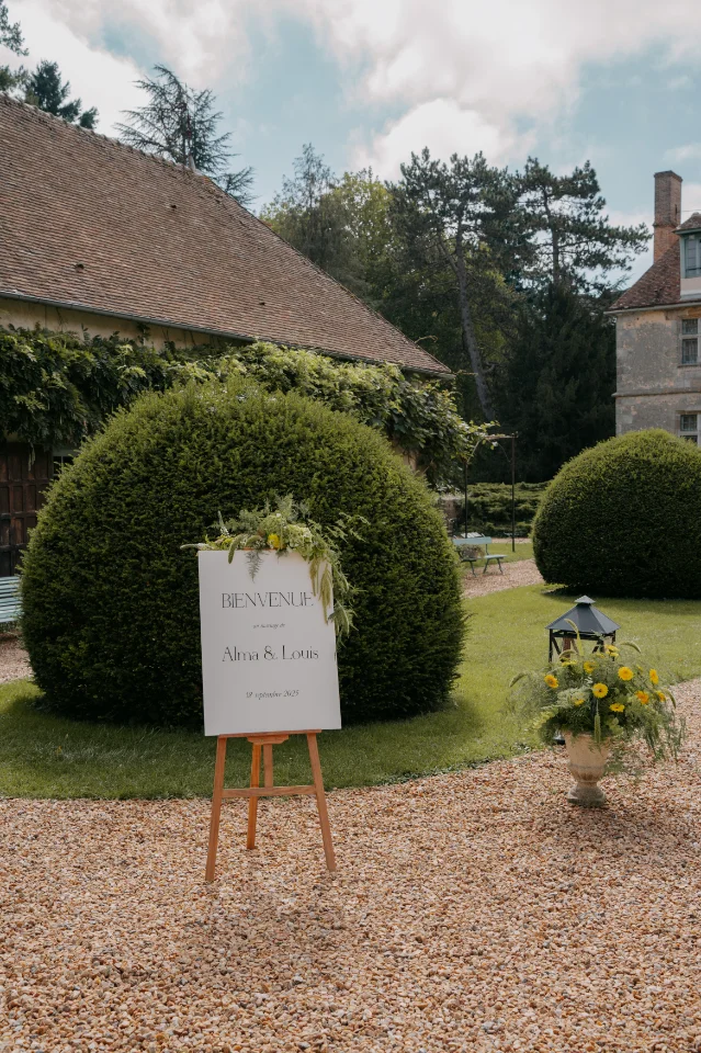Panneau de bienvenue floral pour le mariage d'Alma et Louis dans un jardin champêtre devant un grand buis taillé.