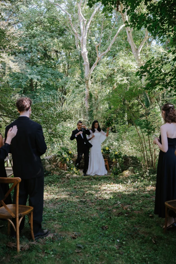 Une mariée en robe blanche et un photographe posent dans un jardin luxuriant, entourés d'invités en tenue noire.