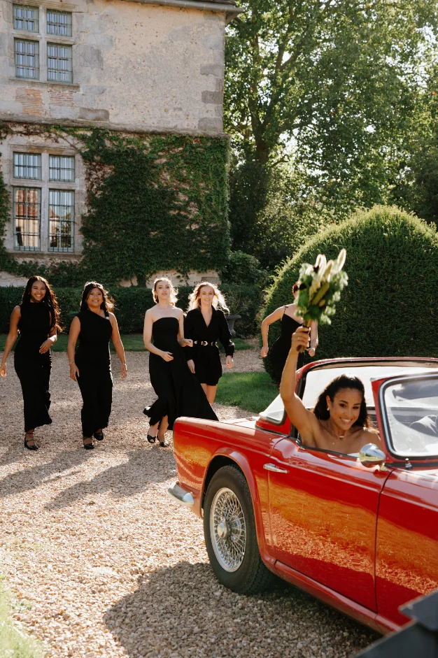 Une mariée dans une voiture rouge vintage salue ses demoiselles d'honneur en robes noires devant un château.