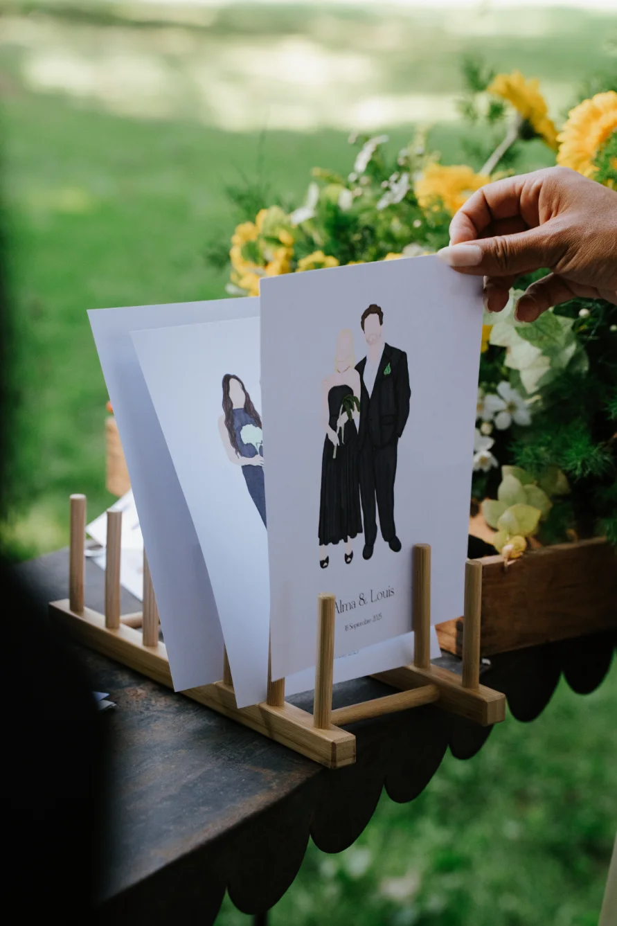 Main tenant des illustrations minimalistes de couples en noir et blanc sur un support en bois, entourées de fleurs.
