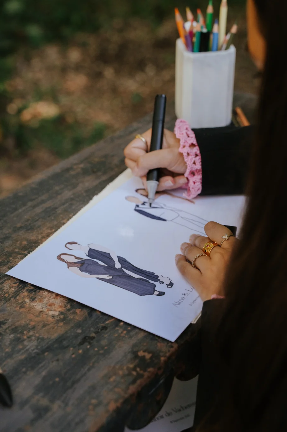 Une artiste dessine des illustrations de couples sur papier, entourée de crayons colorés sur une table en bois rustique.