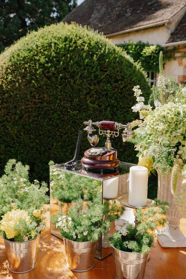 Décoration romantique avec un téléphone ancien sur miroir, entouré de fleurs blanches et jaunes dans des seaux argentés.