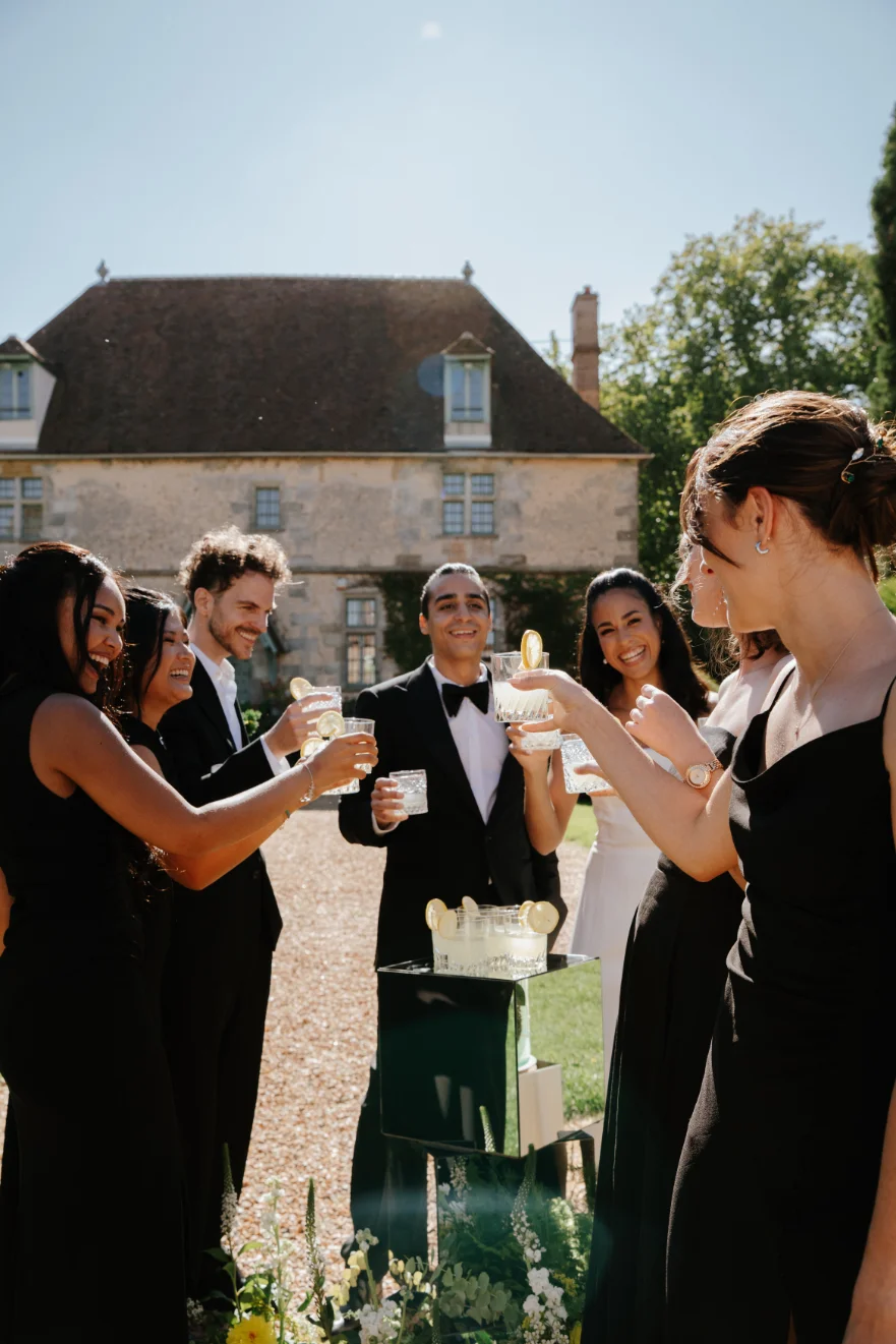 Groupe d'invités élégants en noir et blanc trinquant avec des cocktails devant un manoir ancien et ensoleillé.