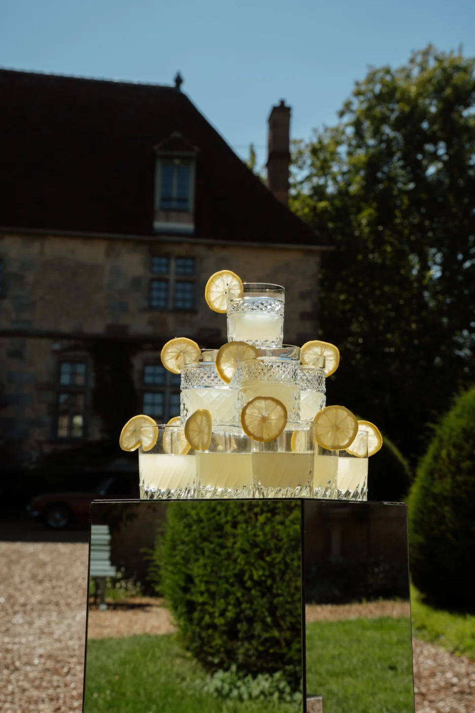 Pyramide de cocktails citronnés décorés de tranches de citron sur un socle miroir, devant un manoir ancien.