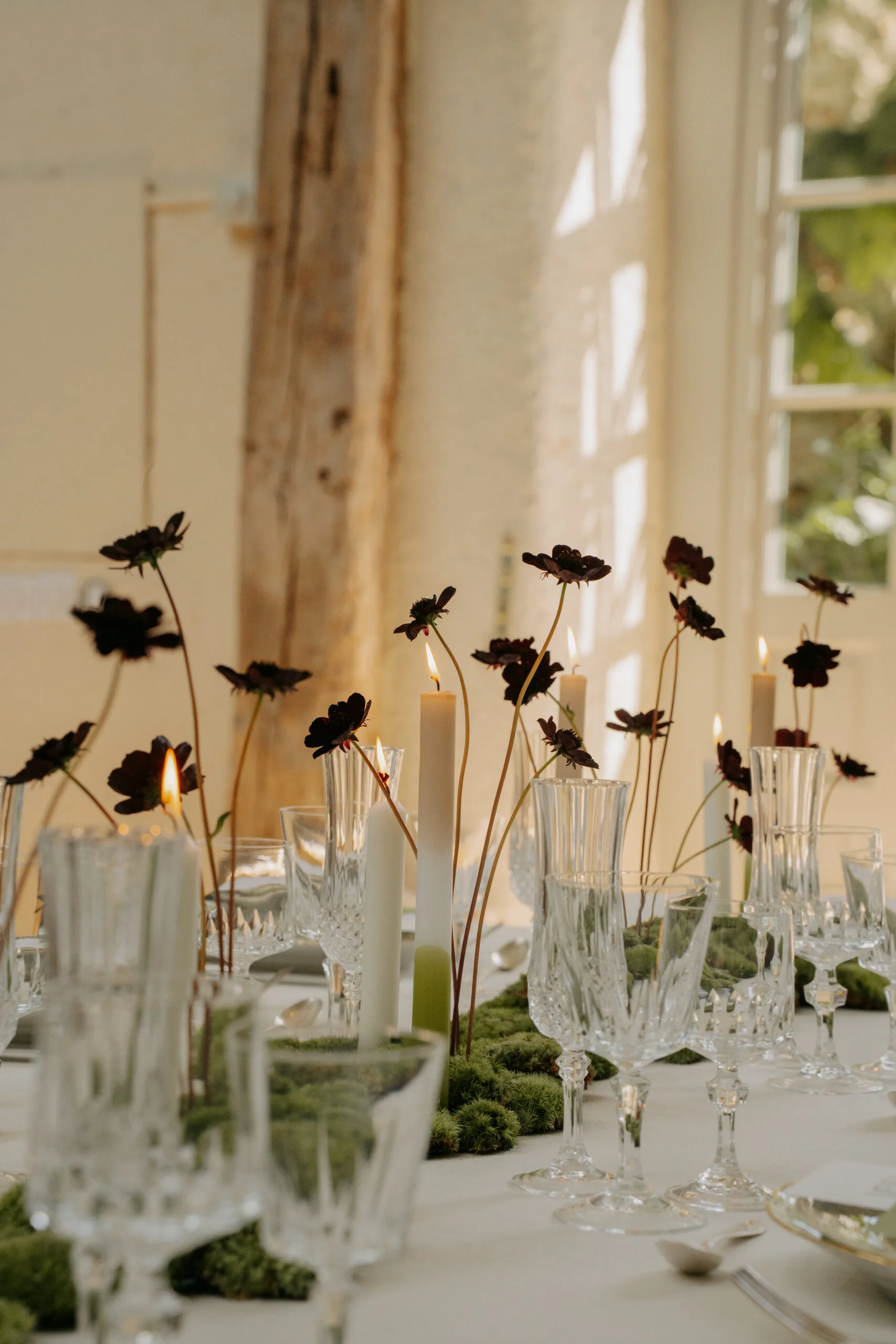 Décoration de table minimaliste avec fleurs sombres, bougies blanches et verres en cristal sur un fond lumineux.