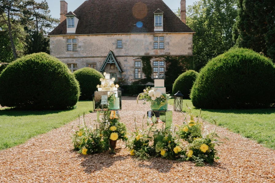 Décoration florale jaune et blanche et gâteaux modernes devant un manoir rustique et des buis taillés.