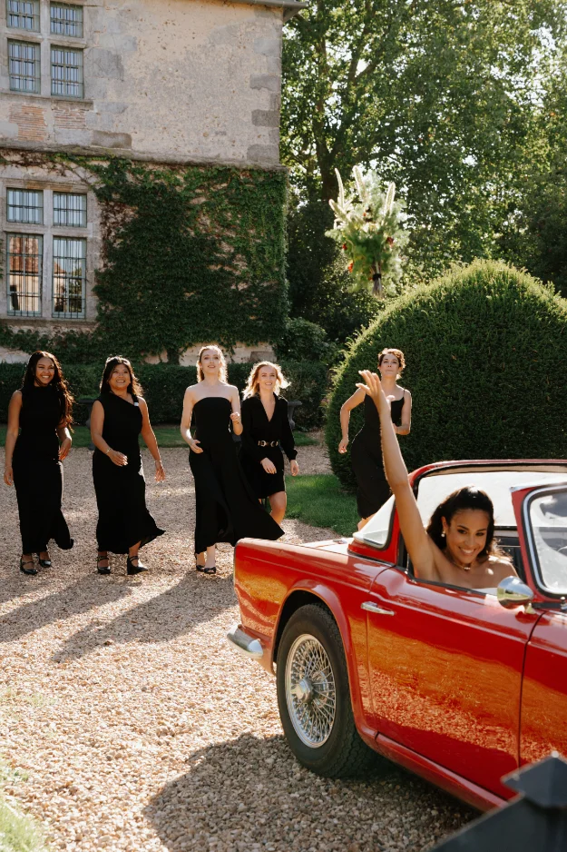 Quatre demoiselles d'honneur en robes noires sourient et s'approchent d'une voiture de collection rouge décapotable.