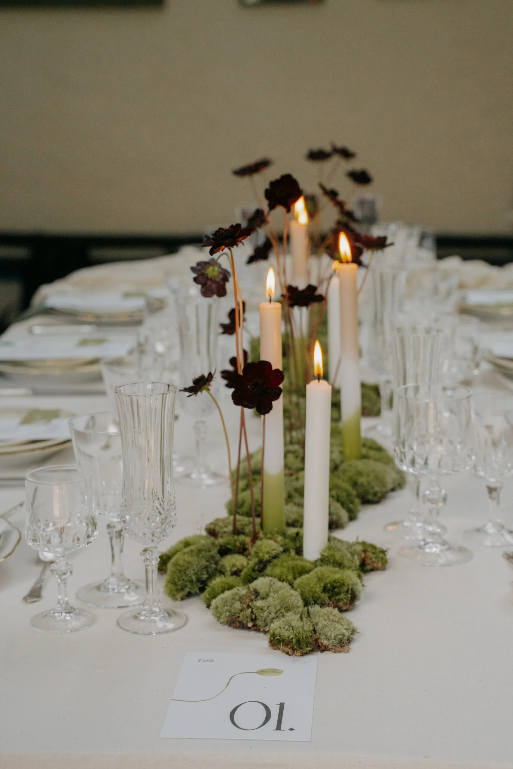 Décoration de table minimaliste avec bougies blanches, mousse verte et fleurs pourpres sur une nappe blanche.