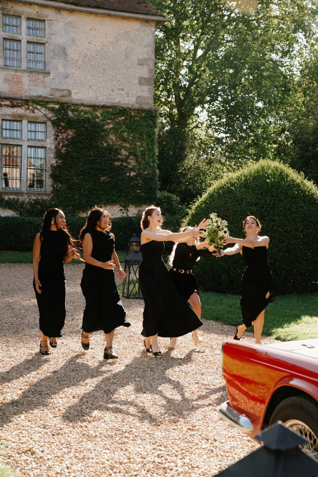 Quatre demoiselles d'honneur en robes noires rient en lançant un bouquet sur un chemin de gravier devant un manoir.