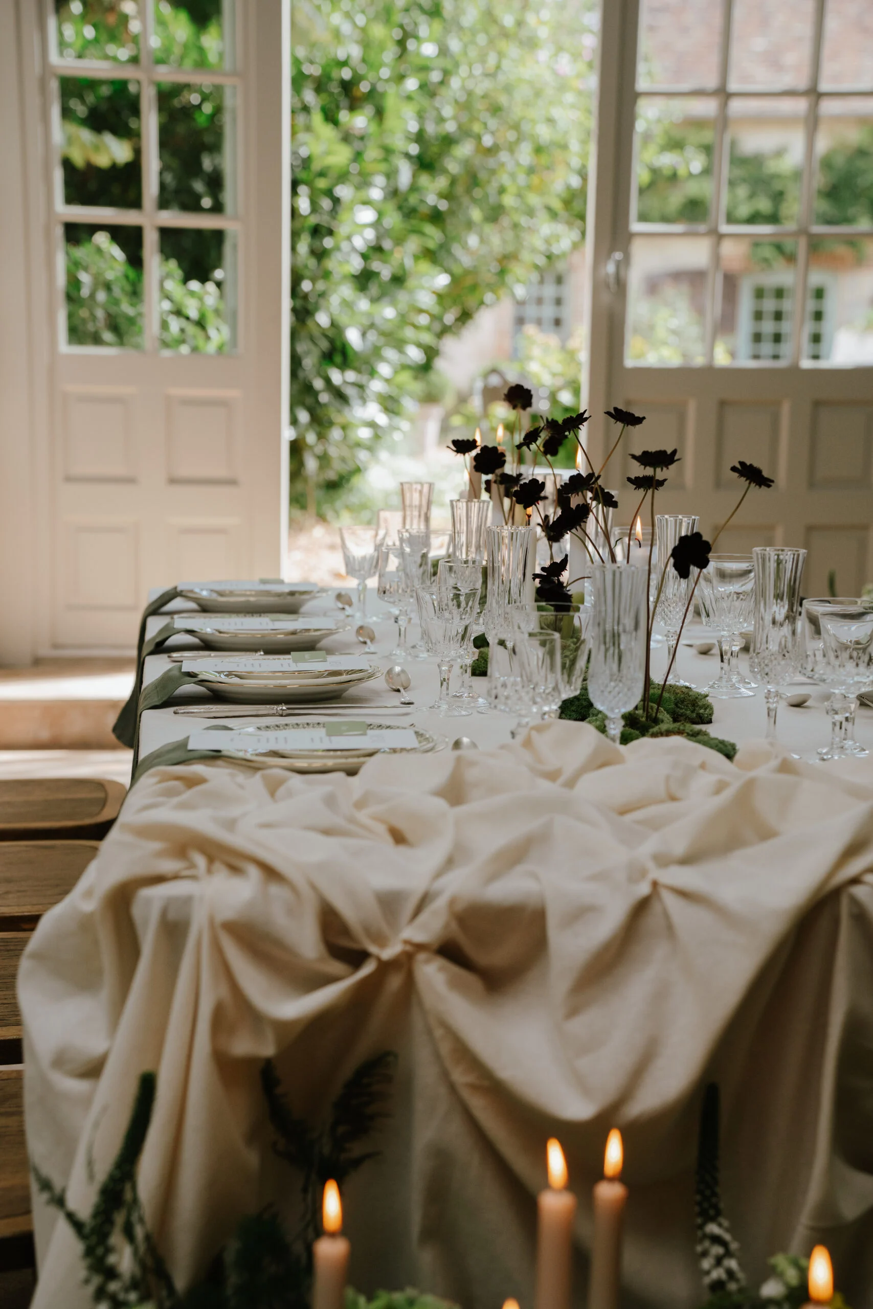 Table élégante dressée avec nappe crème, verres en cristal et fleurs sombres devant des portes-fenêtres lumineuses.