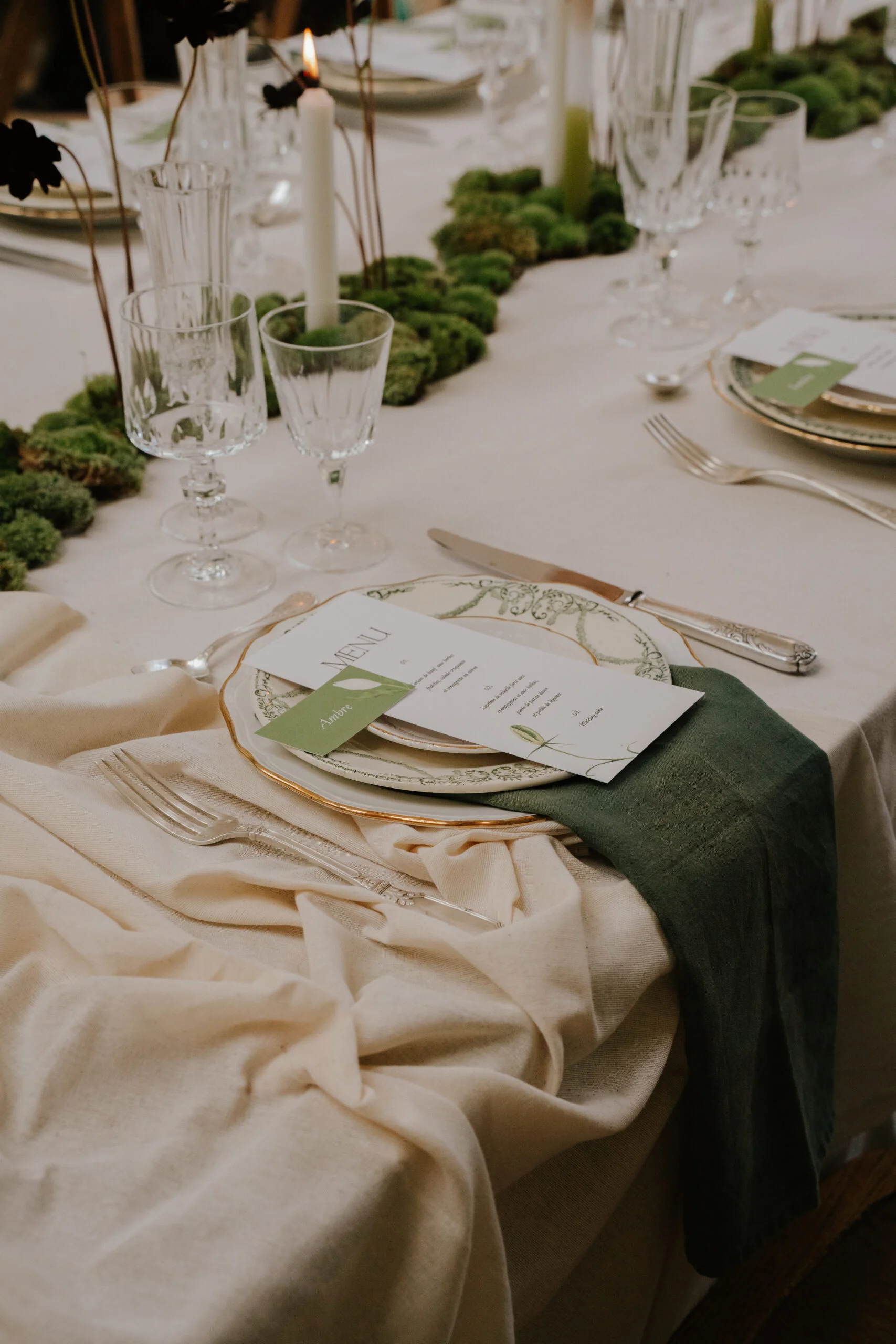 Table élégante avec nappe crème, chemin de table en mousse verte, vaisselle raffinée et menu minimaliste.