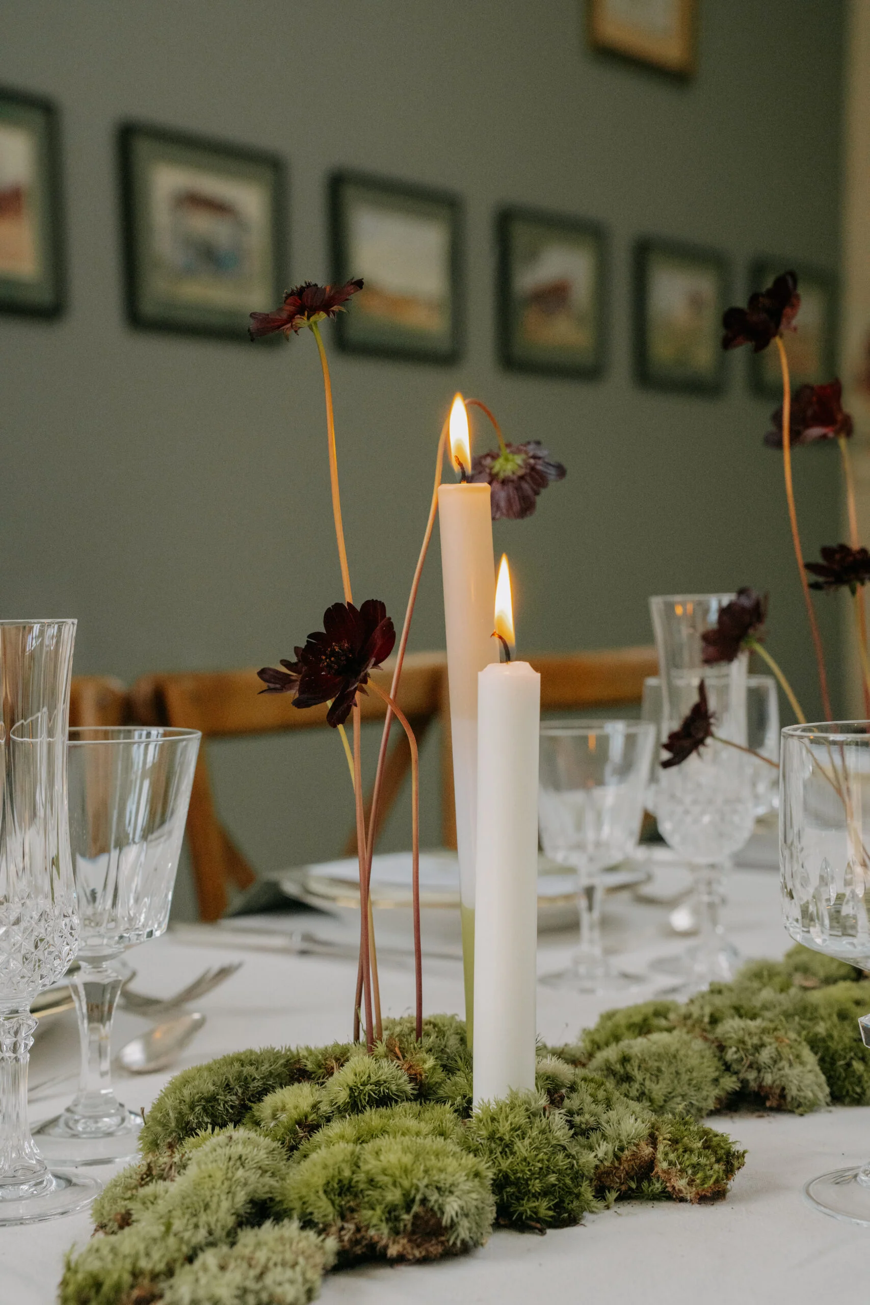 Décoration de table minimaliste avec mousse verte, bougies blanches et fleurs pourpres sur fond vert olive.