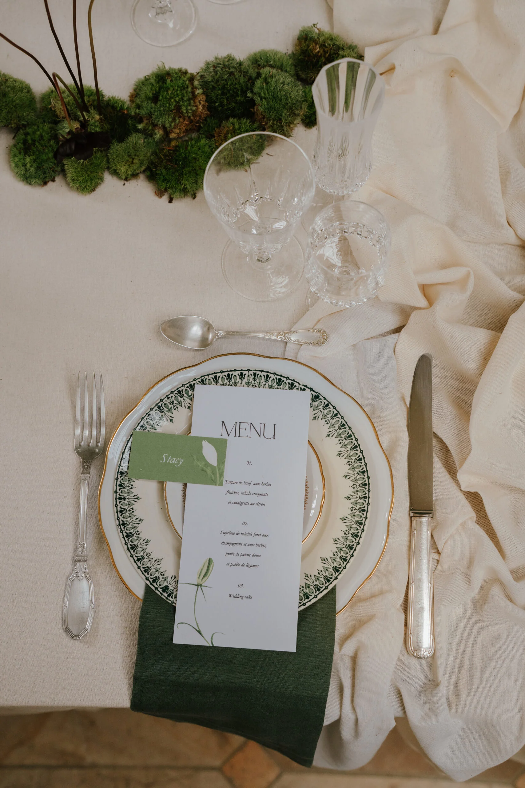 Dressage de table élégant avec menu, serviette vert forêt et mousse végétale sur un nappage crème.