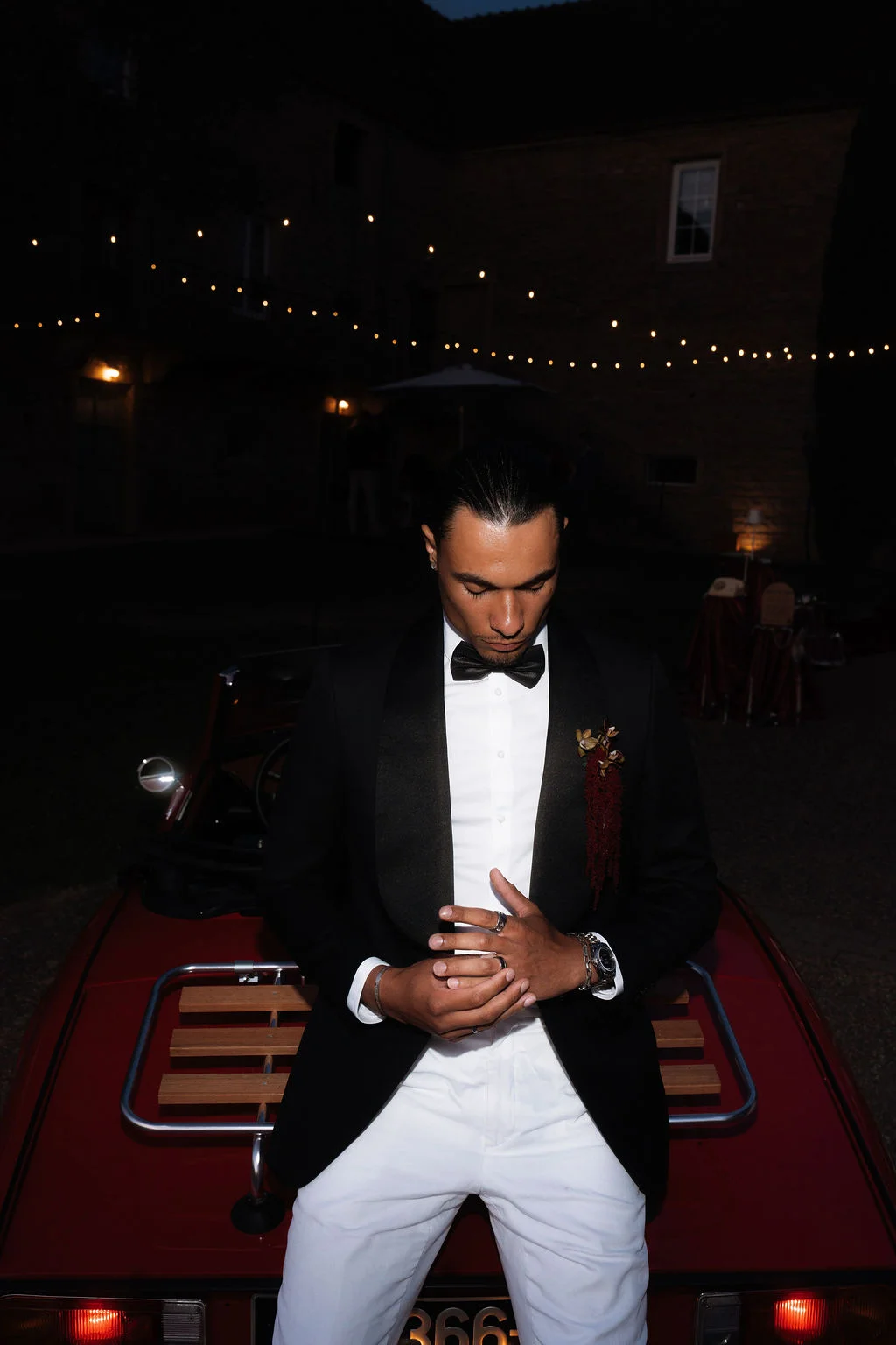 Homme en smoking noir et pantalon blanc posant sur une voiture rouge vintage sous des guirlandes lumineuses nocturnes.