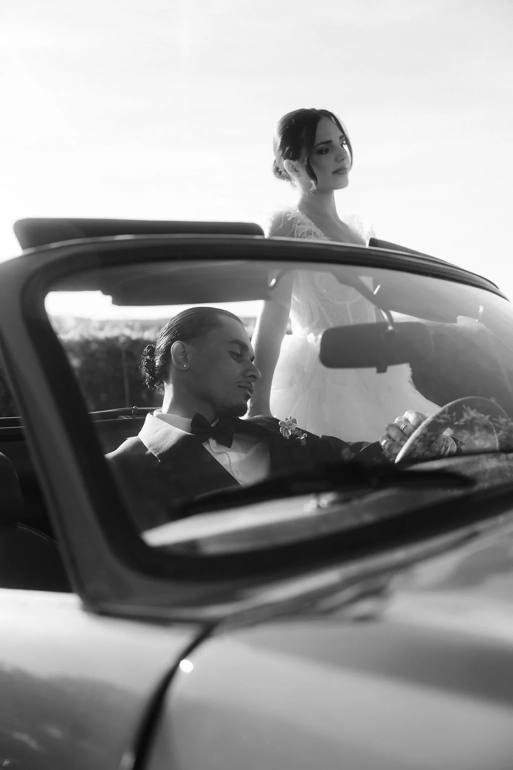 Couple de mariés élégants en noir et blanc, l'homme au volant d'une décapotable et la femme debout à côté.