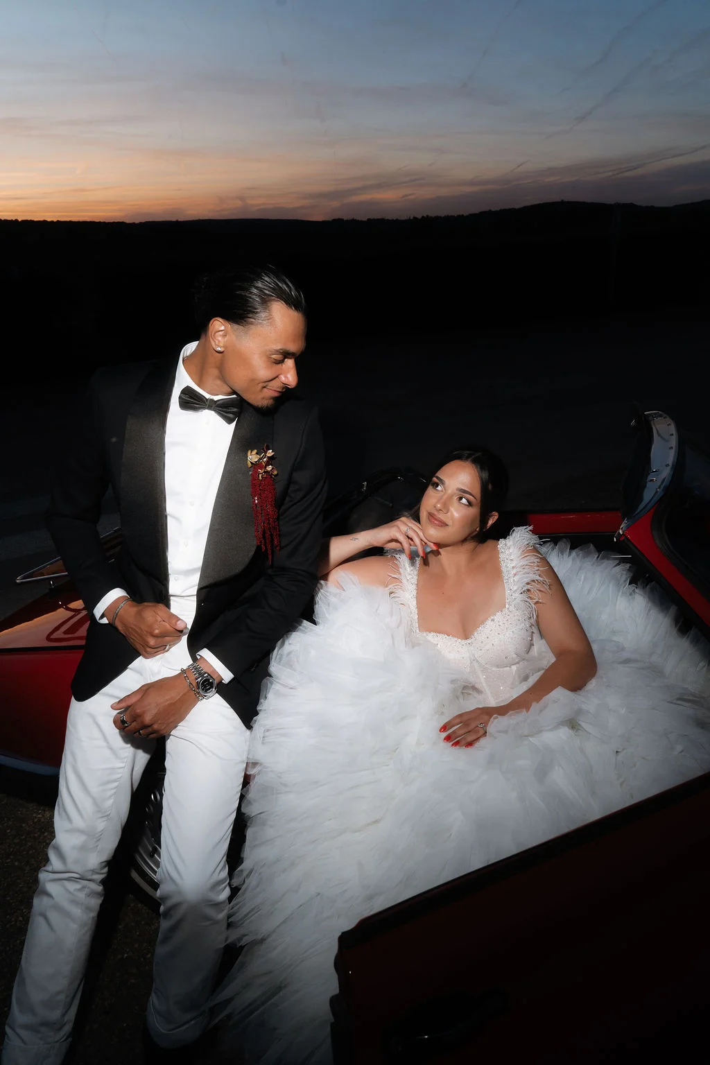 Couple élégant en tenue de mariage posant près d'une voiture rouge vintage au crépuscule.