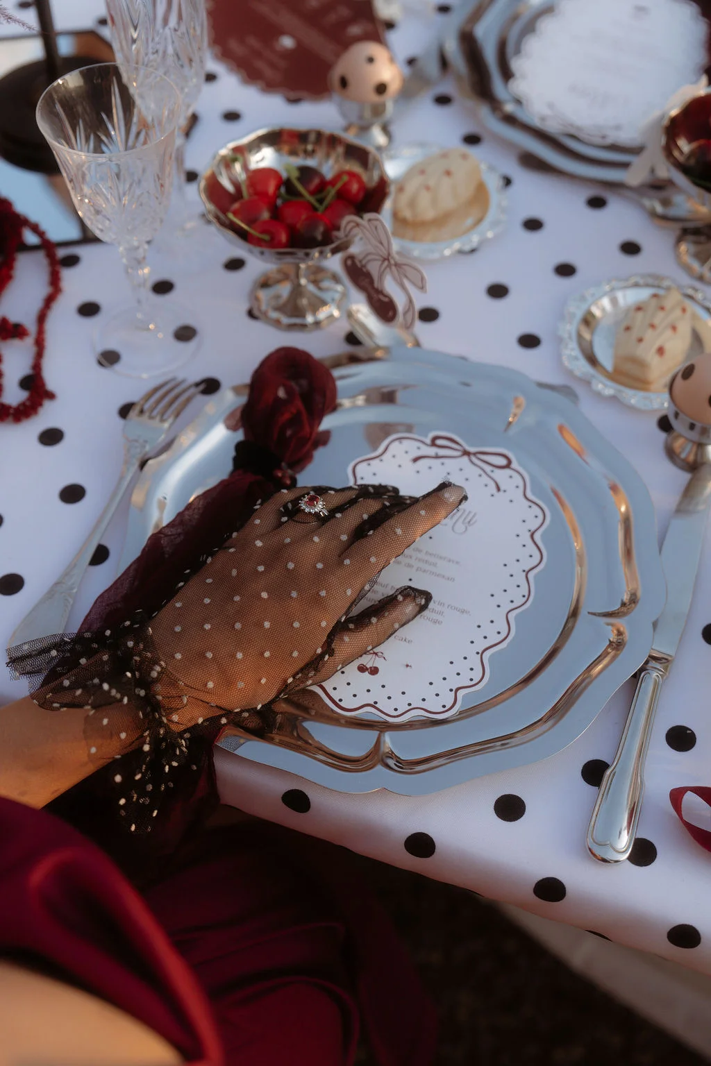 Main gantée de dentelle posée sur une assiette argentée, table blanche à pois noirs avec cerises et touches bordeaux.