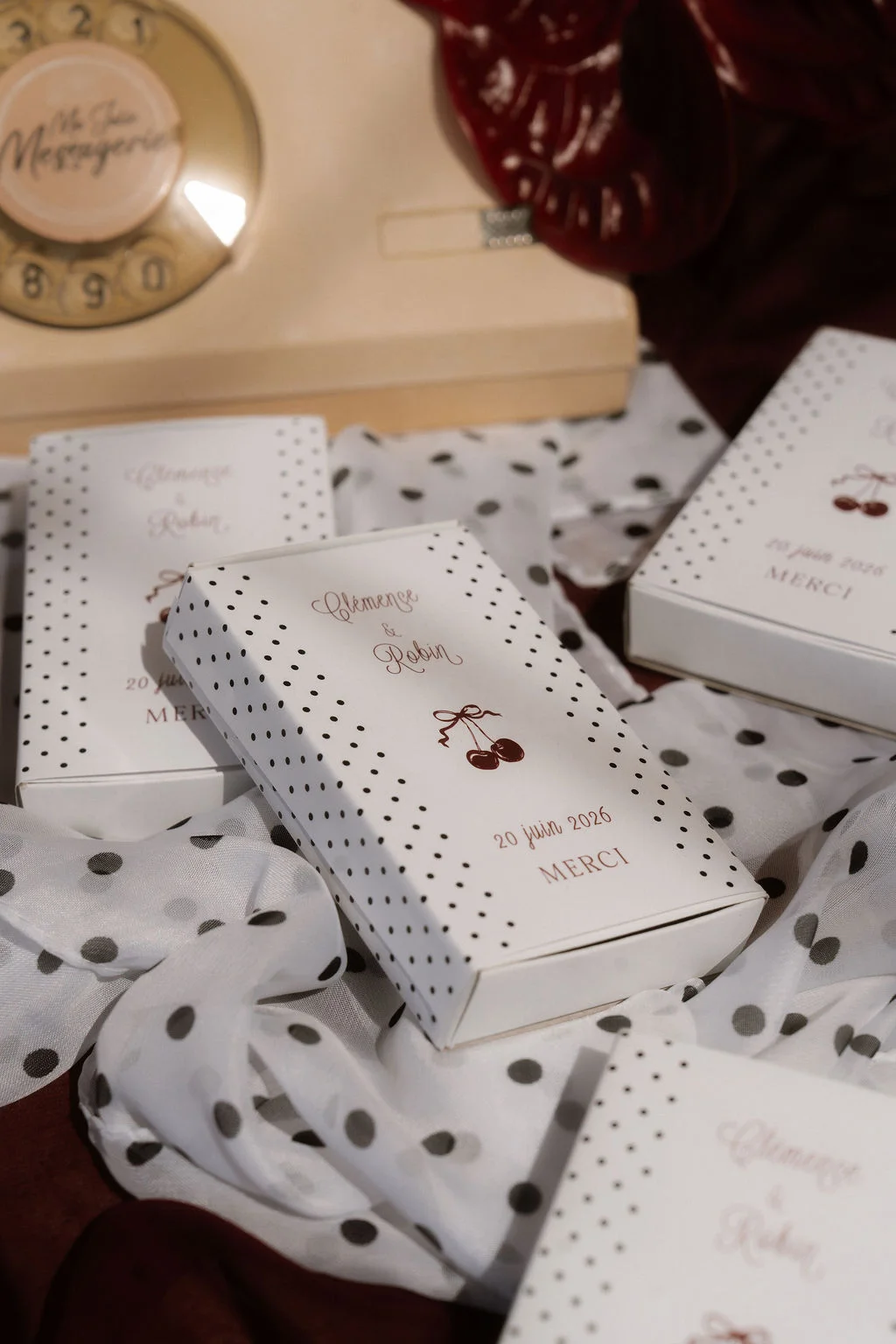 Boîtes de dragées blanches à pois et motifs cerises sur un tissu vaporeux, ambiance romantique et rétro.