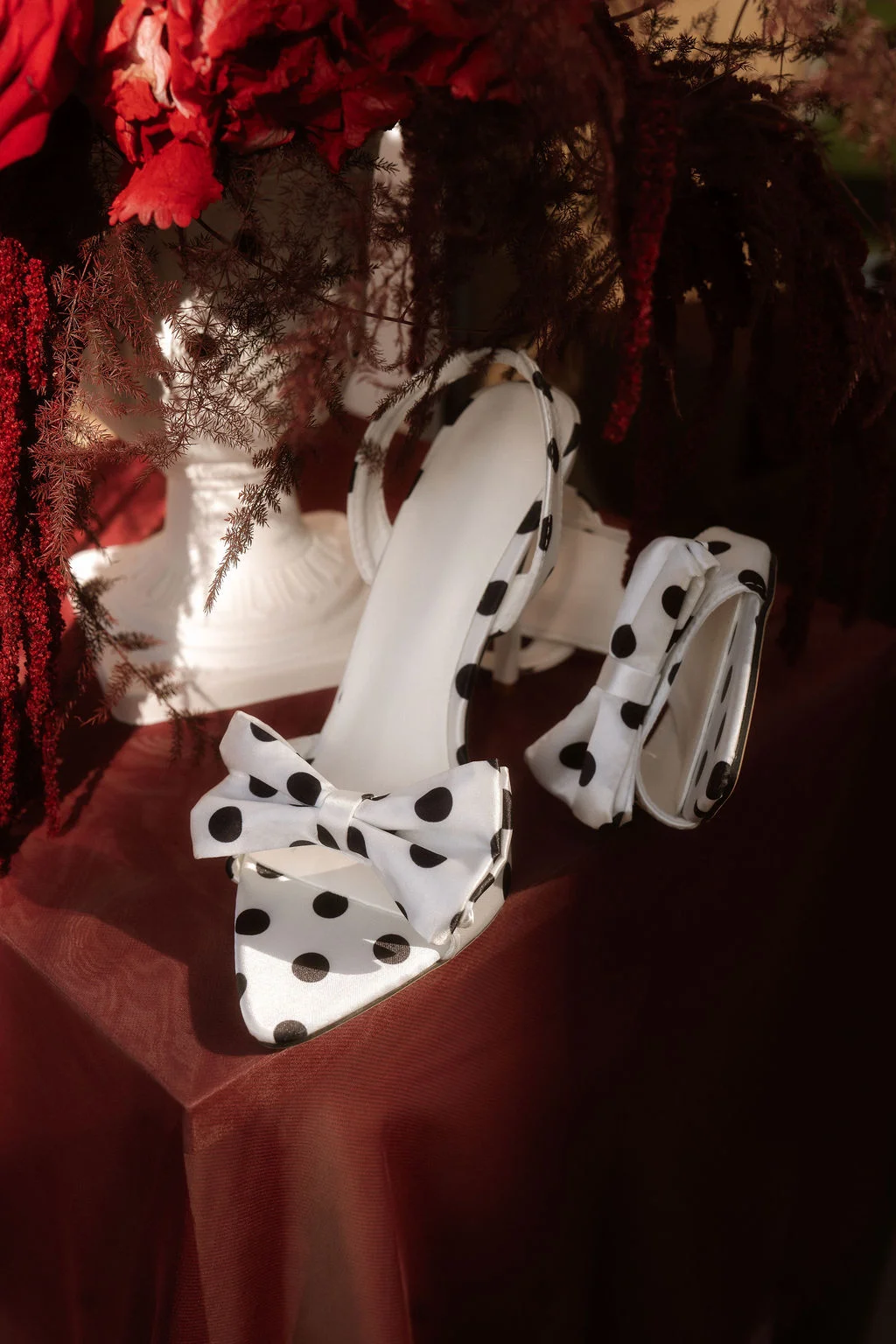 Sandales blanches à pois et nœud posées sur un drapé rouge, entourées de fleurs et de feuillages sombres.