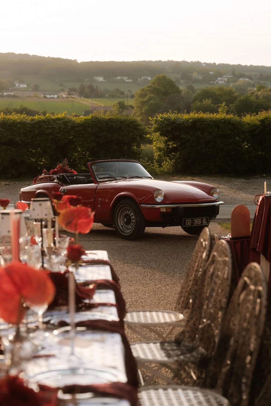 Cabriolet vintage rouge garé près d'une table de réception élégante, sous une lumière dorée de fin de journée.