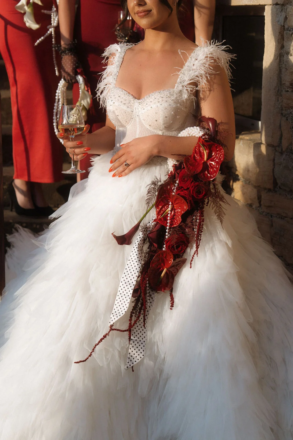 Mariée en robe blanche vaporeuse à plumes, tenant un verre et un bouquet de fleurs rouge cerise et pois.