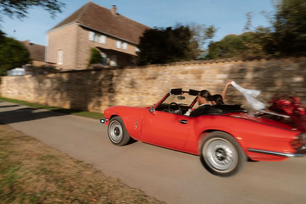 Jeunes mariés dans une décapotable rouge vintage roulant devant un mur de pierre et une maison ancienne.
