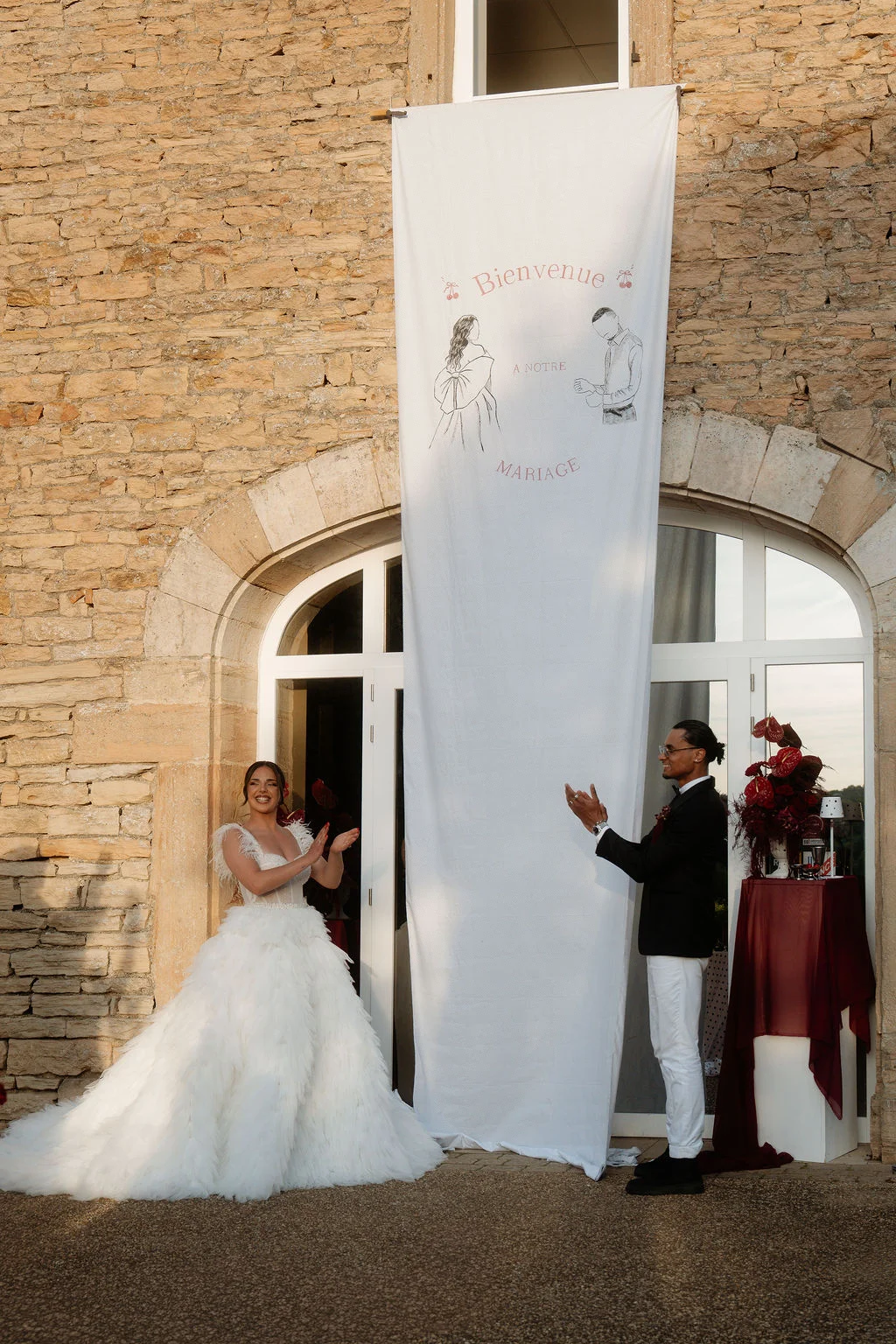 Jeunes mariés s'applaudissant devant un mur en pierre et une bannière blanche lors d'un mariage élégant.