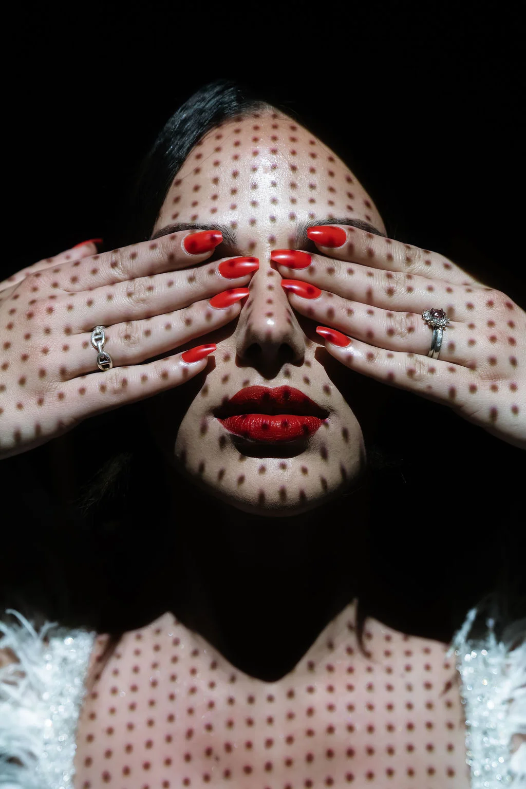 Portrait artistique d'une femme aux points rouges sur la peau, mains sur les yeux et lèvres rouge vif sur fond noir.