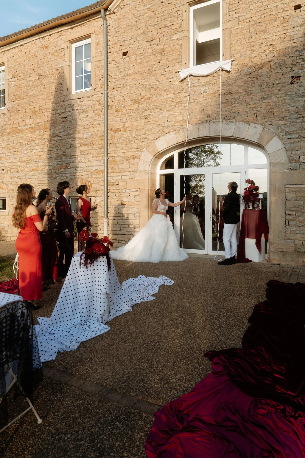 Une mariée en blanc et ses demoiselles d'honneur en rouge posent devant un bâtiment en pierre sous une lumière dorée.