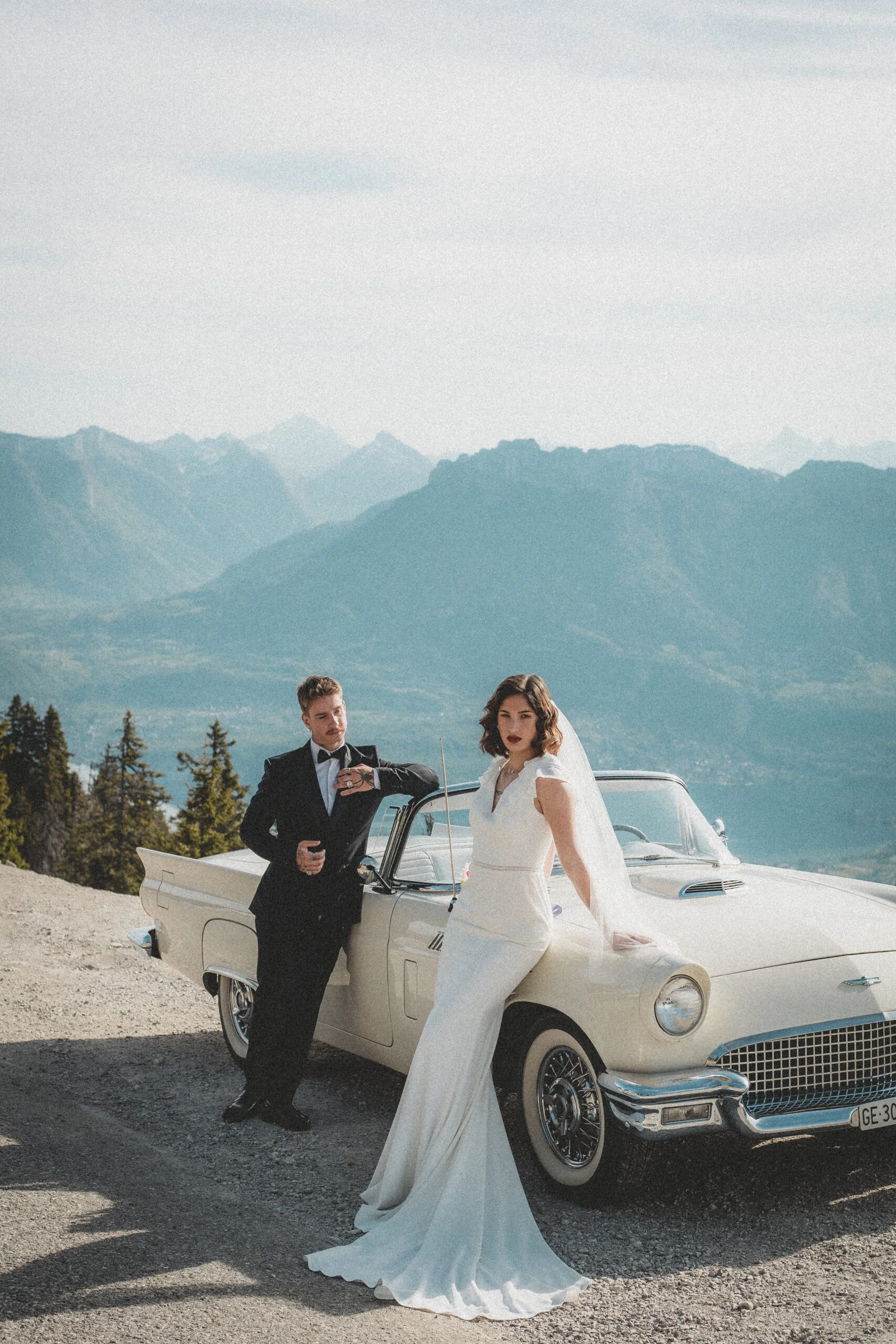 Couple marié élégant posant près d'une voiture vintage blanche devant un panorama montagneux bleuté.