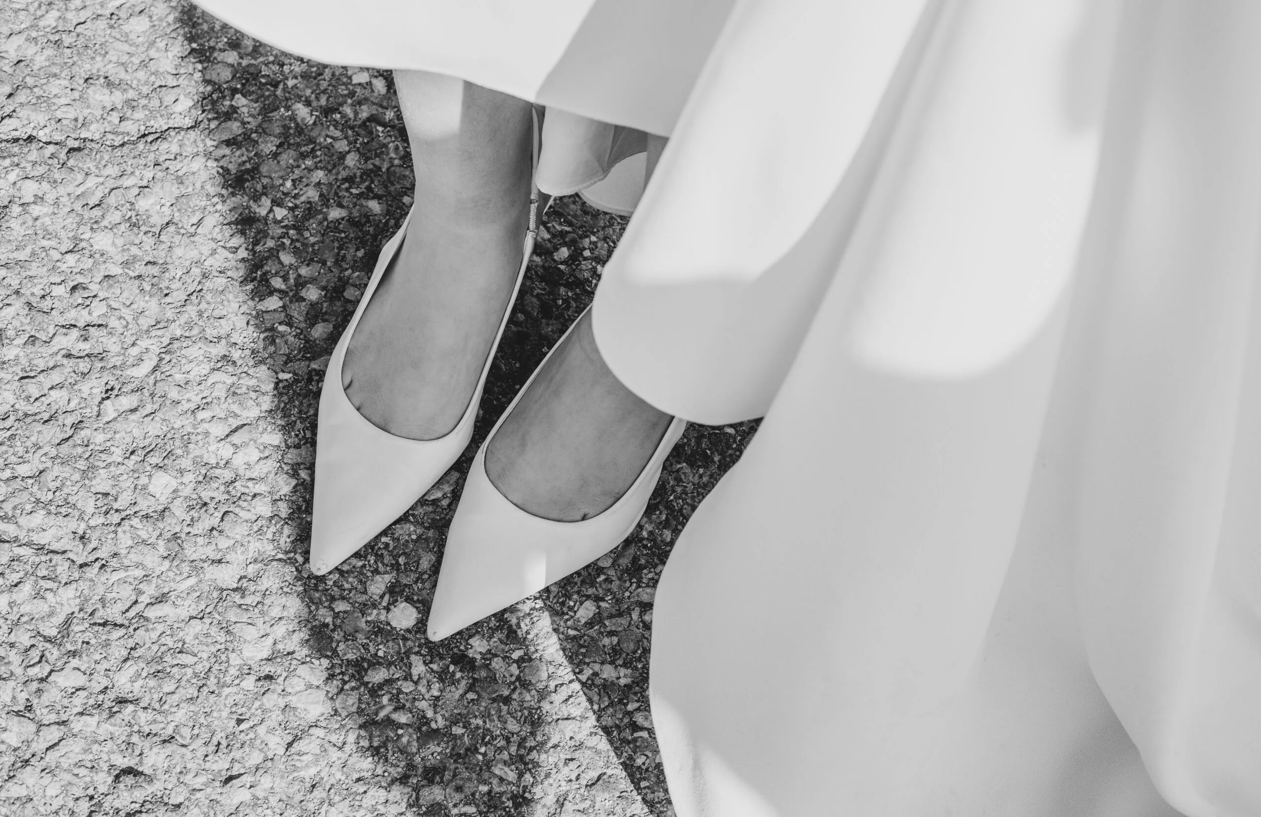 Gros plan en noir et blanc des pieds d'une mariée portant des escarpins pointus sous une robe fluide.