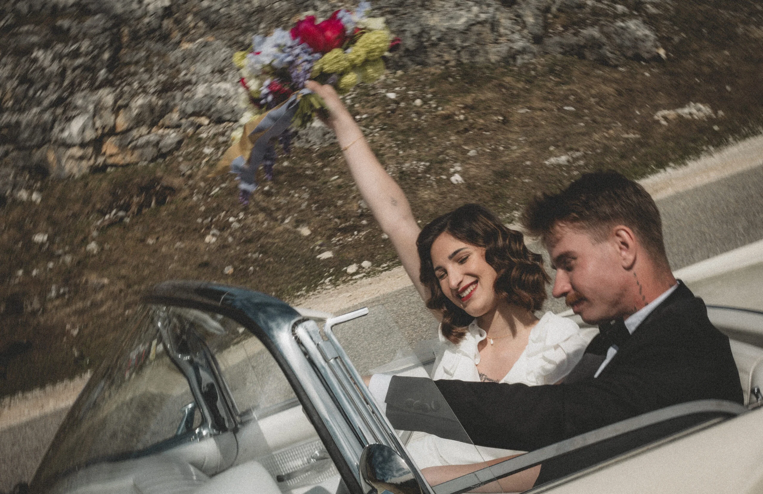 Jeunes mariés dans une voiture décapotable vintage, la mariée brandissant un bouquet coloré dans une ambiance rétro.