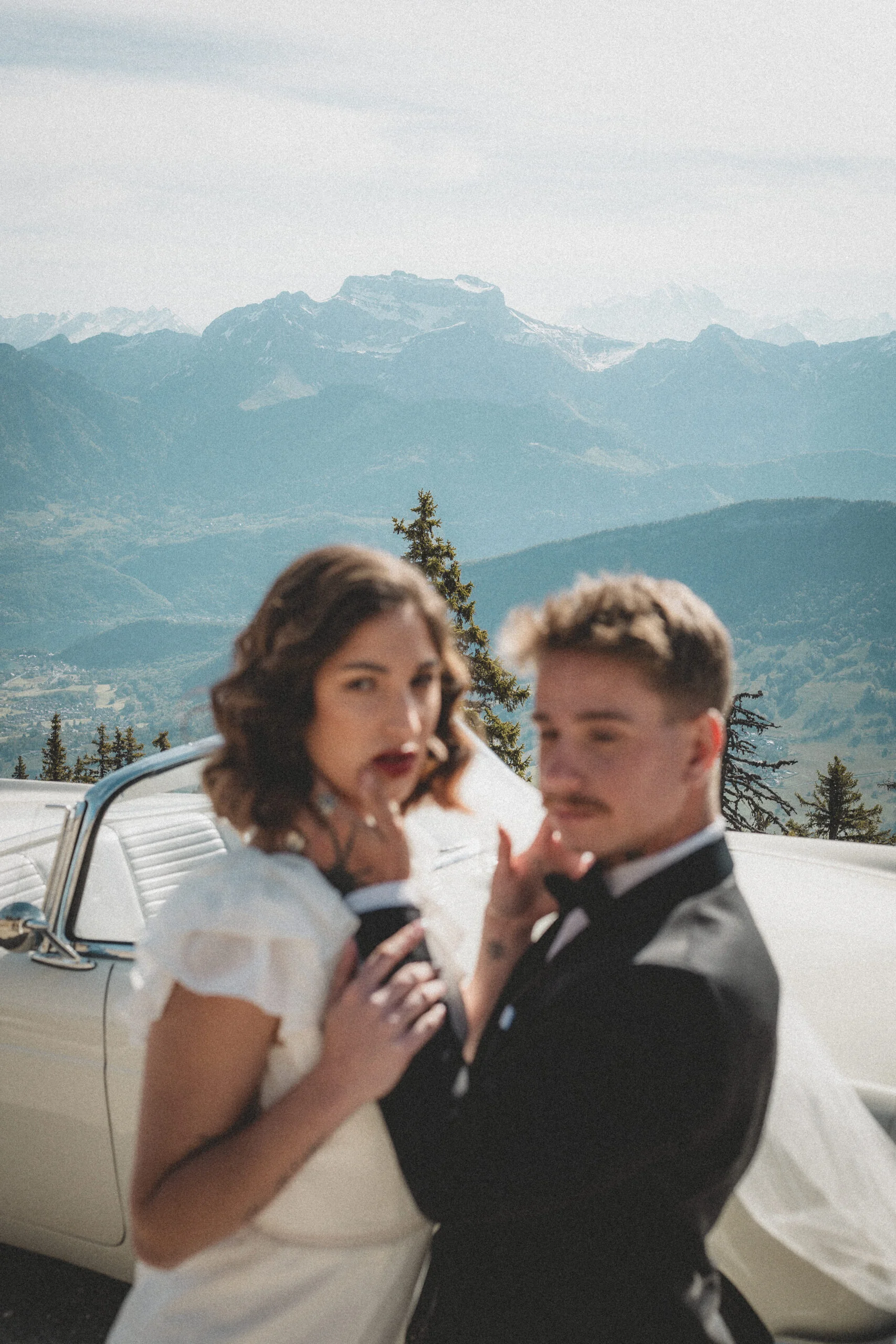 Couple de mariés au style rétro posant devant une voiture blanche avec des montagnes bleutées en arrière-plan.