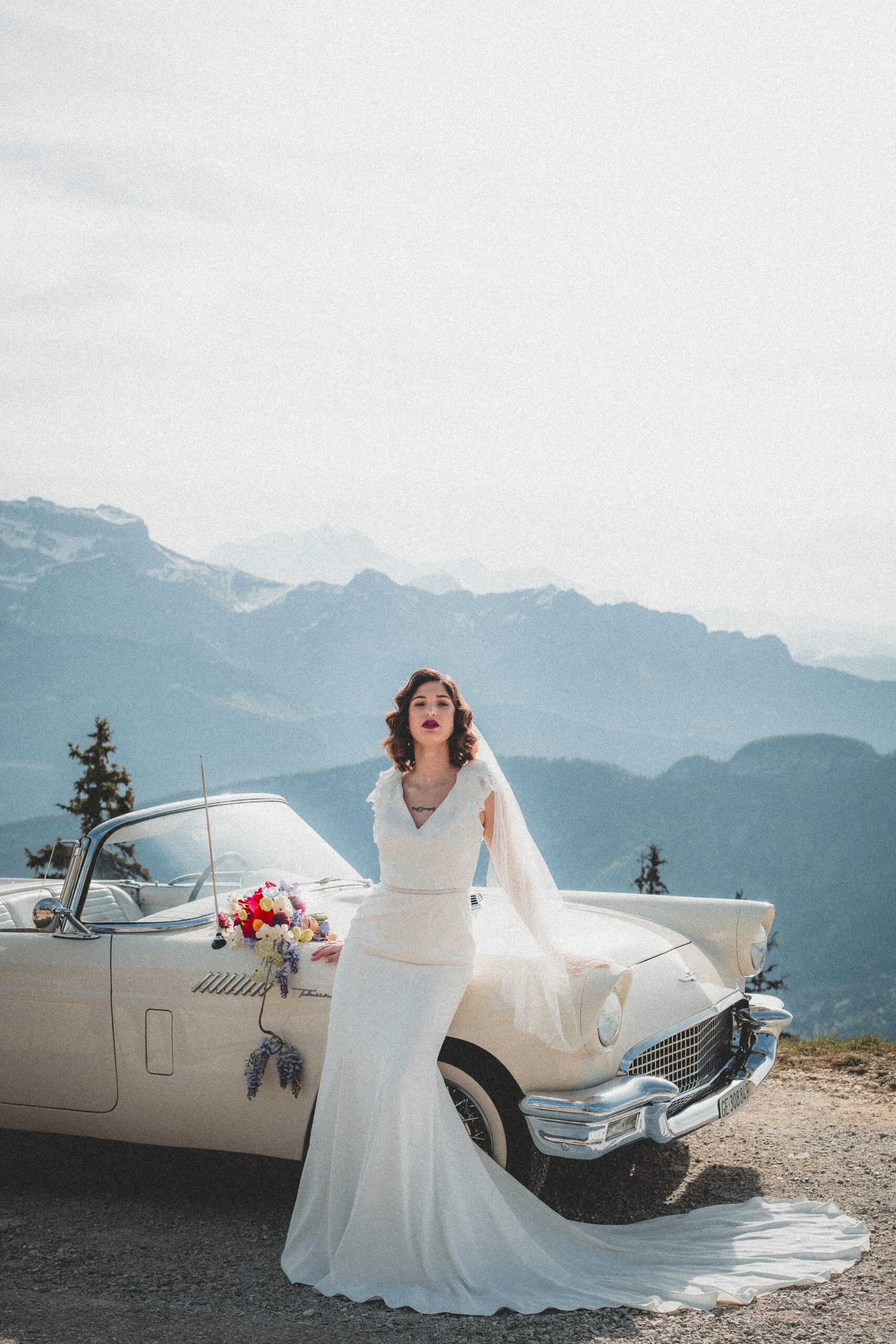 Mariée en robe blanche et voile posant avec un bouquet coloré devant une voiture ancienne, face aux montagnes.