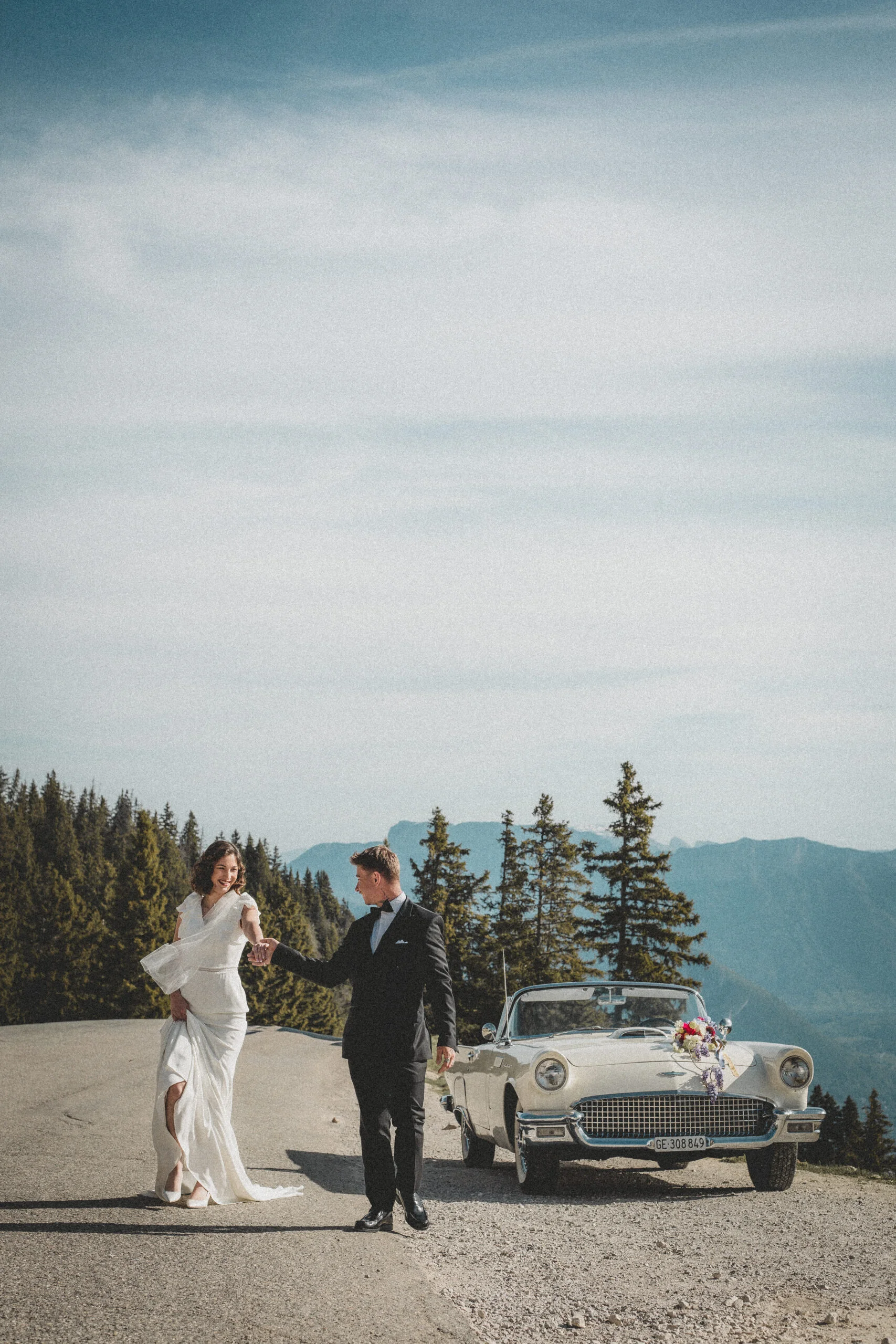 Jeunes mariés élégants main dans la main devant une voiture vintage blanche, dans un paysage montagneux et boisé.