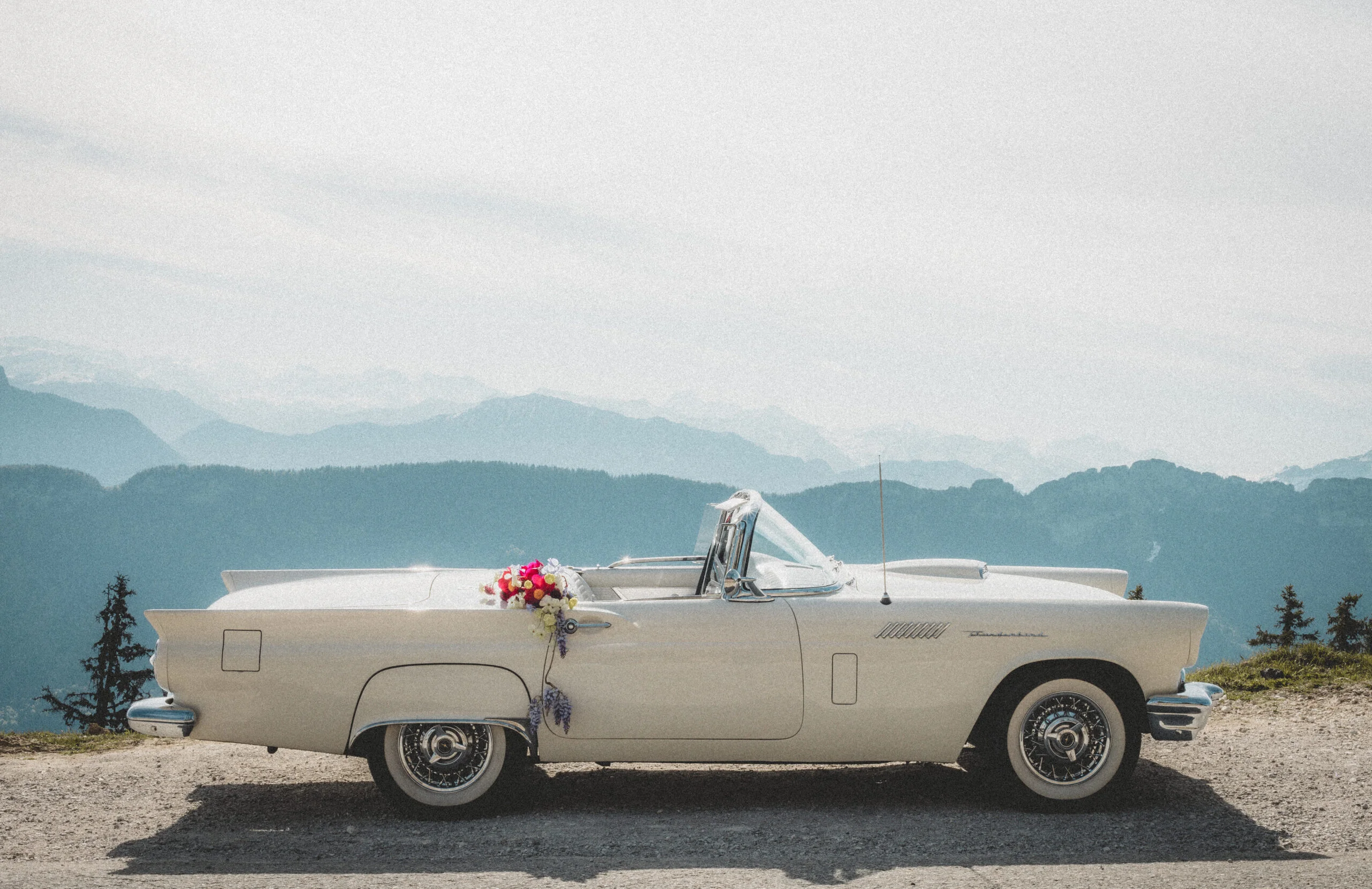 Voiture de collection blanche décorée de fleurs, garée face aux montagnes sous un ciel clair.