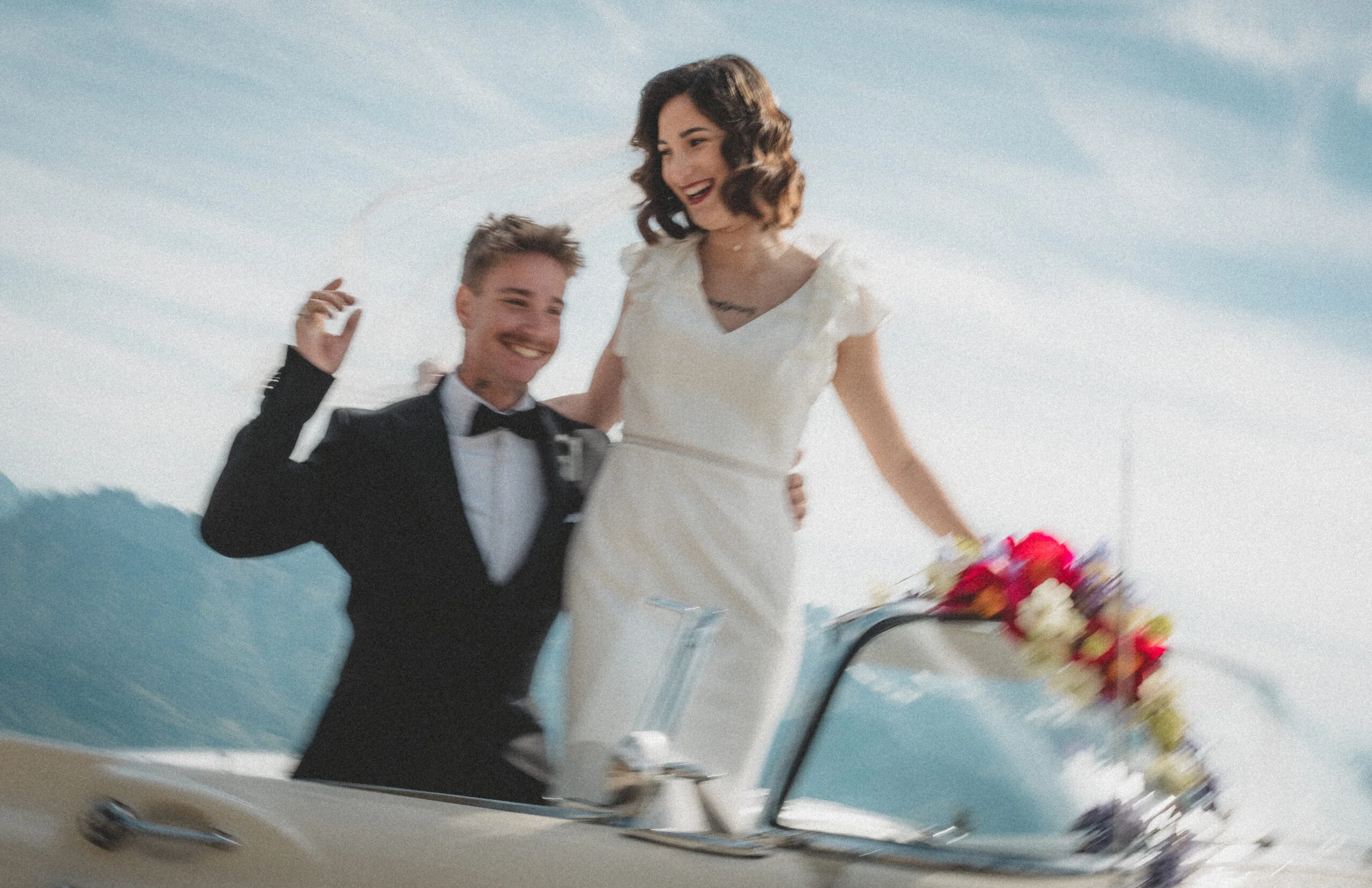 Jeunes mariés souriants dans une décapotable vintage avec des fleurs, ambiance rétro et romantique.