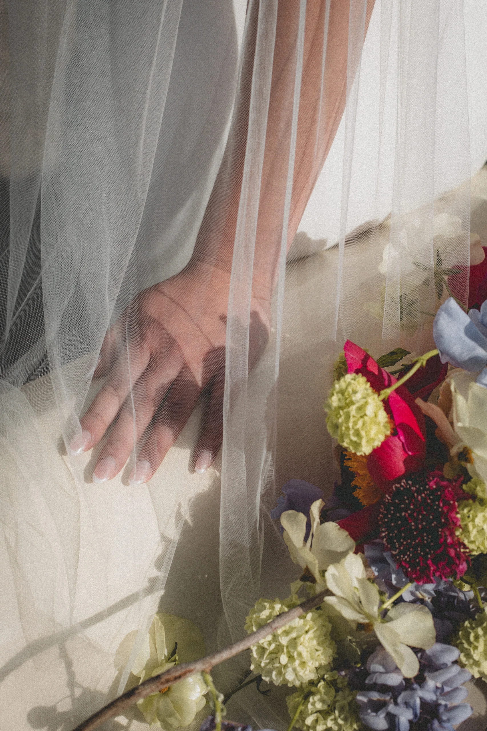 Main de mariée sous un voile blanc vaporeux à côté d'un bouquet coloré, ambiance romantique et rétro.