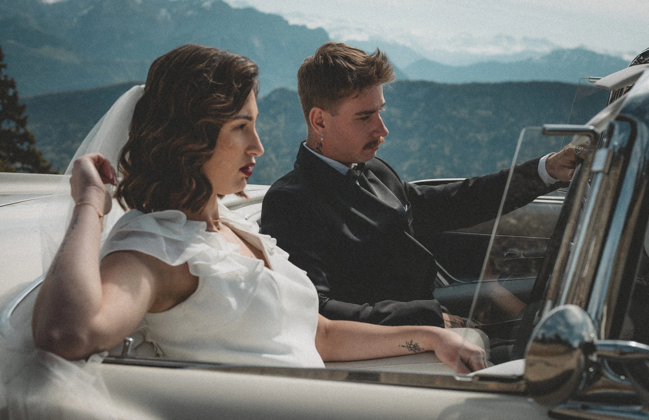 Jeunes mariés élégants dans une voiture ancienne, posant devant un paysage montagneux dans une ambiance rétro.