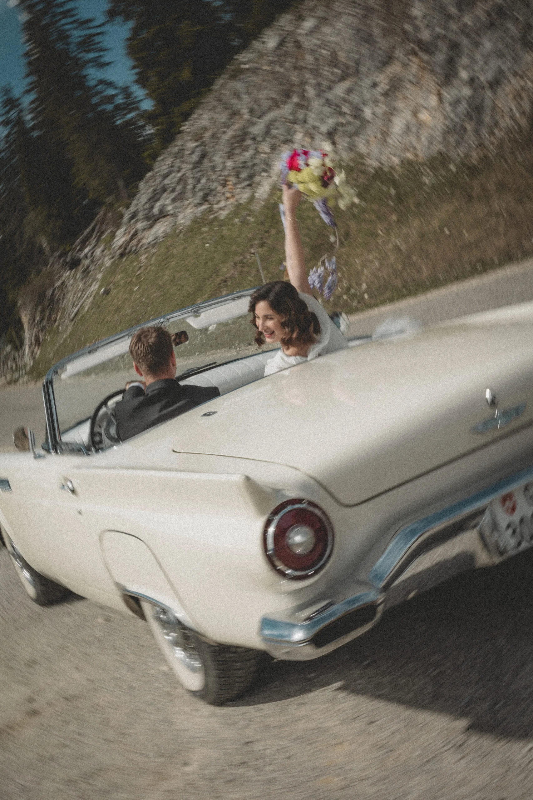 Une mariée joyeuse brandit son bouquet depuis une décapotable blanche vintage sur une route de montagne.