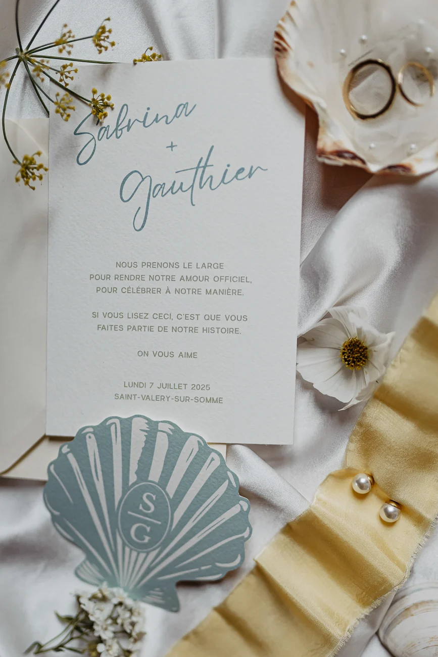 Faire-part de mariage blanc et bleu pastel avec coquillages et alliances sur un tissu satiné blanc et doré.