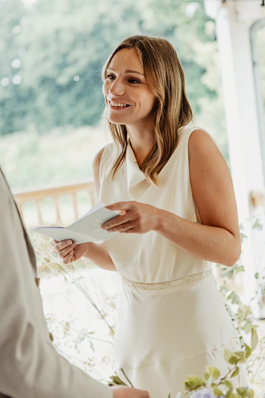 Une mariée souriante en robe blanche lit ses vœux lors d'une cérémonie intime et lumineuse en extérieur.