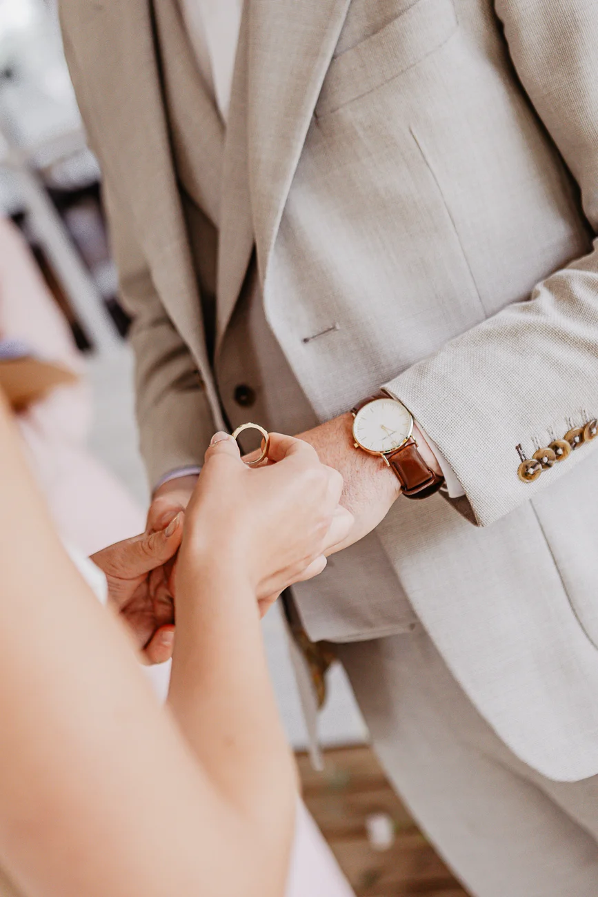 Gros plan sur les mains d'un couple et une alliance, le marié portant un costume beige et une montre en cuir marron.