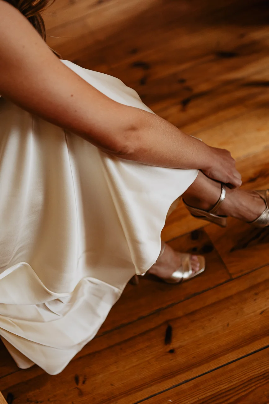 Une mariée en robe blanche satinée ajuste ses sandales dorées sur un parquet en bois chaleureux.